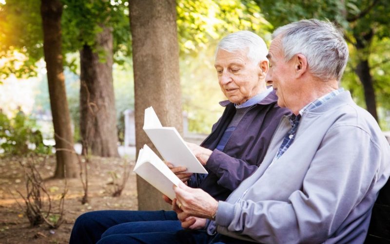 Two senior men reading books in park. One is reading a passage to his amused partner.